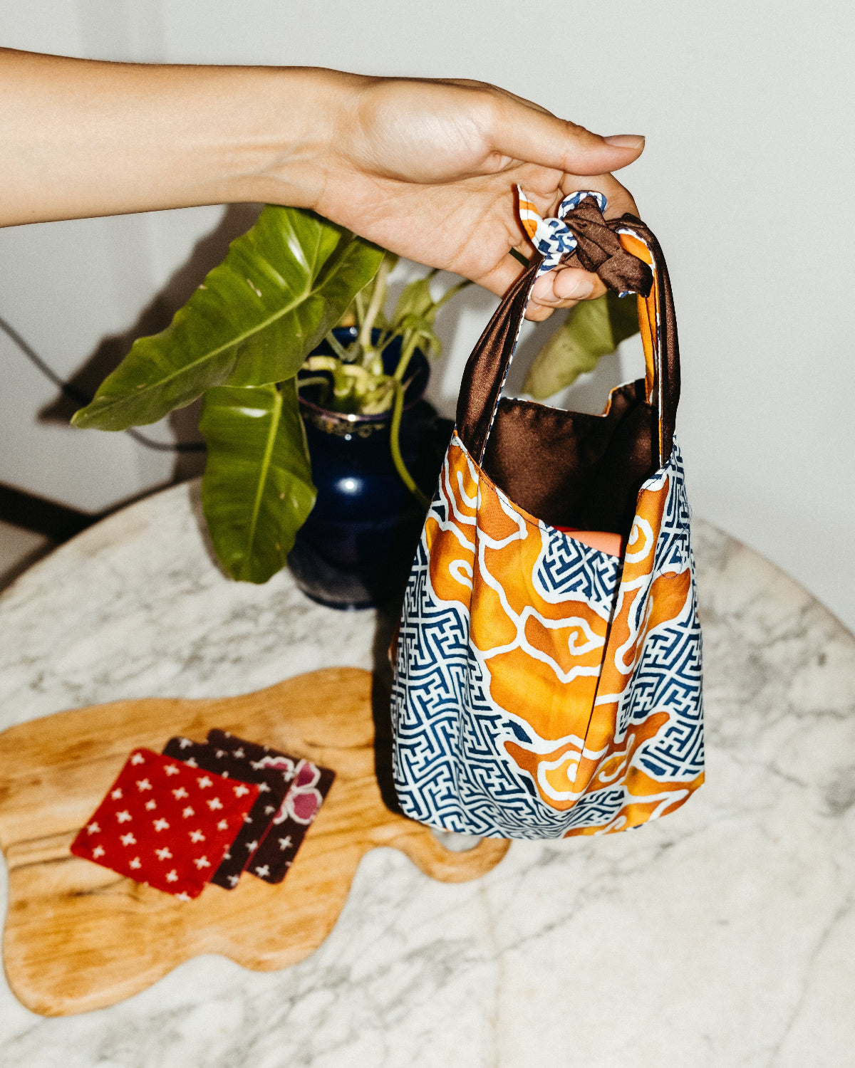 Hand holding a colorful batik patterned bag on a marble surface with a plant in the background